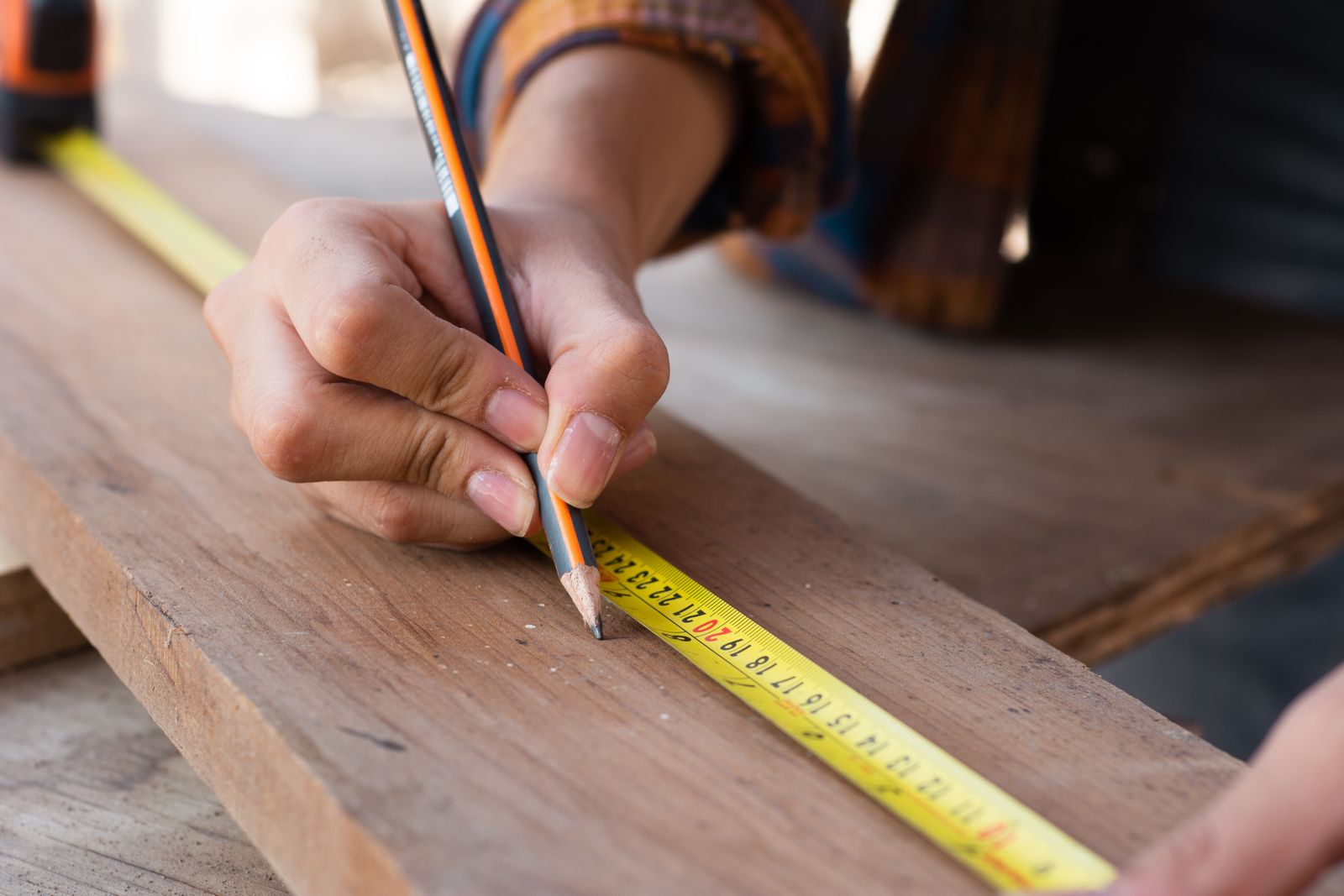 A hand marking a wooden board with a pencil along a yellow tape measure