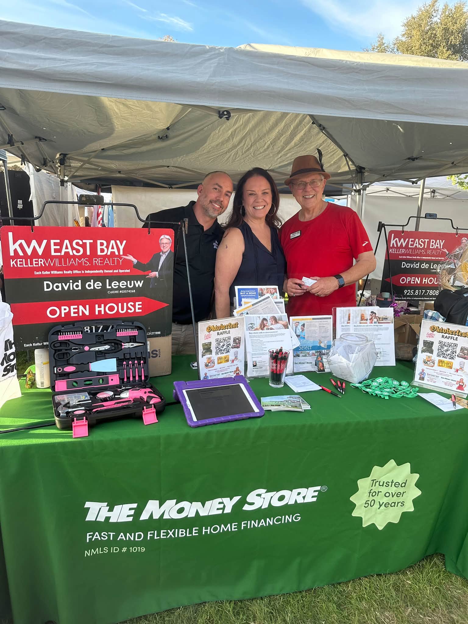 Three people smiling together at a booth at the Oktoberfest in Walnut Creek, with a Keller Williams sign on the left and a green tablecloth on the table
